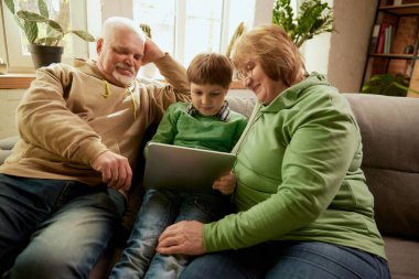 Mutlu dost canlısı aile, büyük anne, büyük baba ve torun kanepede oturup birlikte vakit geçiriyorlar, modern aletler kullanıyorlar, konuşuyorlar, çalışıyorlar. Aşk, ilgi, destek, hobiler ve boş zaman aktiviteleri kavramı