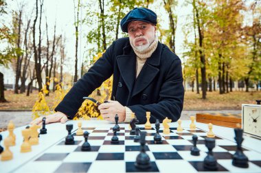 Sonbaharda güpegündüz parkta satranç oynayan son sınıf öğrencisinin portresi. Zor bir strateji. Boş zaman aktivitesi, arkadaşlık, spor, sonbahar sezonu, oyun, eğlence, eski nesil.