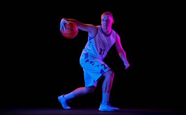 Salya akıtıyor. Genç adam, profesyonel basketbolcu topla hareket halinde siyah arka planda neon ışık filtresiyle izole edilmiş. Spor anlayışı, enerji, yetenek, takım yarışması. Sporcu enerjik görünüyor.