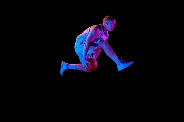 In motion. Active athletic male basketball player jumping with basketball ball isolated over dark background in purple neon light. Concept of energy, professional sport, hobby, competition