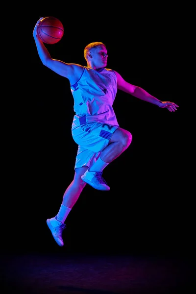 In motion. Active athletic male basketball player jumping with basketball ball isolated over dark background in purple neon light. Concept of energy, professional sport, hobby, competition