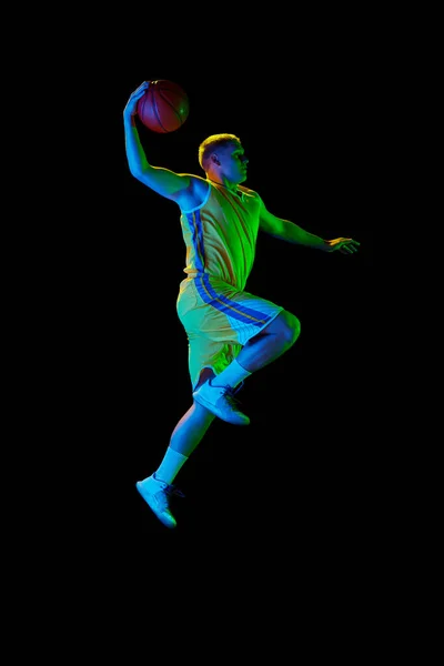 若いアクティブな選手のダイナミックな肖像画 ネオンの光の暗い背景の上に隔離されたボール ジャンプ ドリブルを練習するスポーツの制服を着た男性バスケットボール選手 広告のコピースペース — ストック写真
