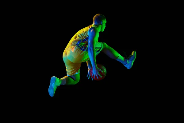 In motion. Active athletic male basketball player jumping with basketball ball isolated over dark background in blue neon light. Concept of energy, professional sport, hobby, competition, achievement.