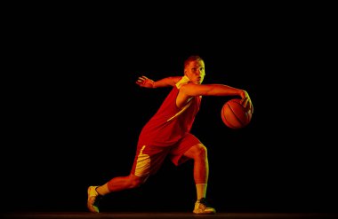 Salya akıtıyor. Genç adam, profesyonel basketbolcu topla hareket halinde siyah arka planda neon ışık filtresiyle izole edilmiş. Spor anlayışı, enerji, yetenek, takım yarışması. Sporcu enerjik görünüyor.