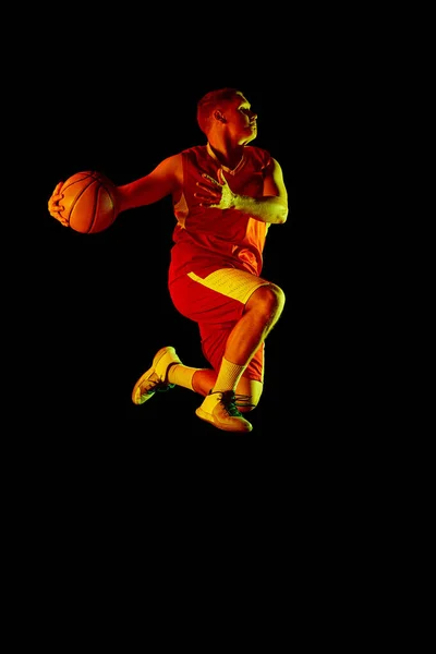 In motion. Active athletic male basketball player jumping with basketball ball isolated over dark background in red neon light. Concept of energy, professional sport, hobby, competition, achievement.