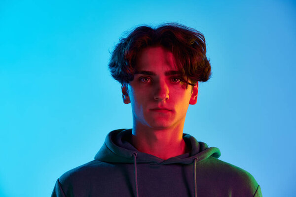 Portrait of young man with curly hair posing, seriously looking at camera isolated over blue background in neon light. Concept of youth culture, emotions, facial expression, fashion. Copy space for ad