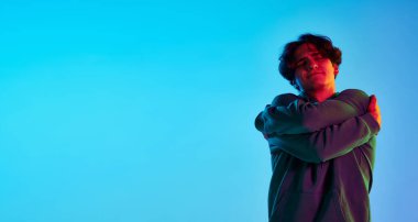 Kıvırcık saçlı genç bir adamın portresi. Mavi arka planda kendine sarılıyor. Kendini sevmek. Psikoloji. Gençlik kültürü kavramı, duygular, yüz ifadesi, moda. El ilanı