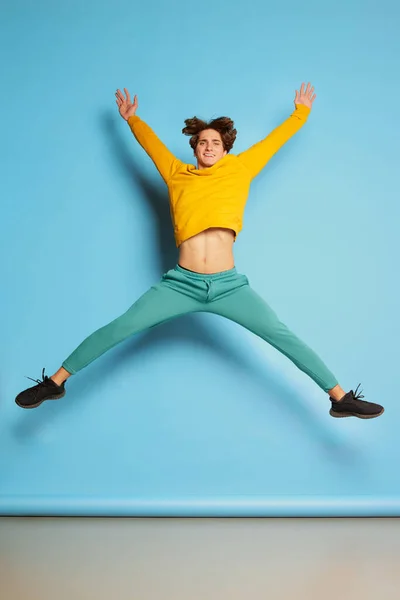 Portrait of young man with curly hair posing, jumping in excitement isolated over blue background. Concept of youth culture, emotions, facial expression, fashion. Copy space for ad