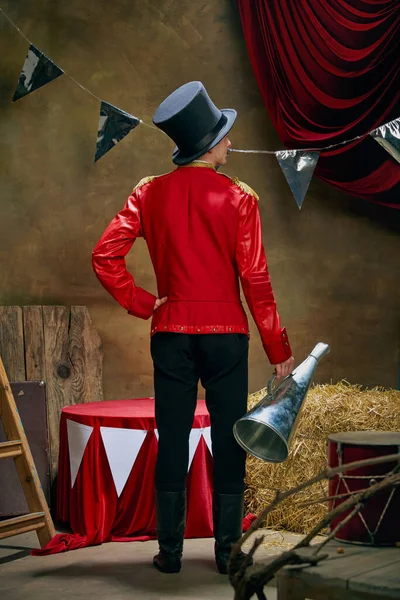 Back view. Illusionist or showman. Young man retro circus entertainer announces start of show isolated over dark retro circus backstage background. Concept of emotions, art, sales