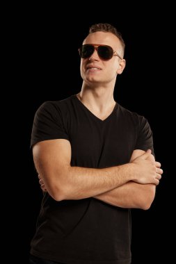 Siyah stüdyo arkaplanından izole edilmiş güneş gözlüğü takan koyu renk tişörtlü yakışıklı bir adam portresi. Erkek sağlığı, yaşam tarzı, moda, duygular, refah kavramı. İlan için alanı kopyala