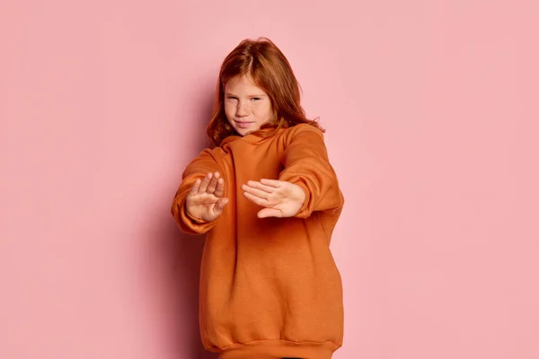 Stop sign. One scared little girl, kid with long red hair and freckles posing isolated over pink background. Concept of children emotions, casual sports fashion style, facial expressions. Copy space