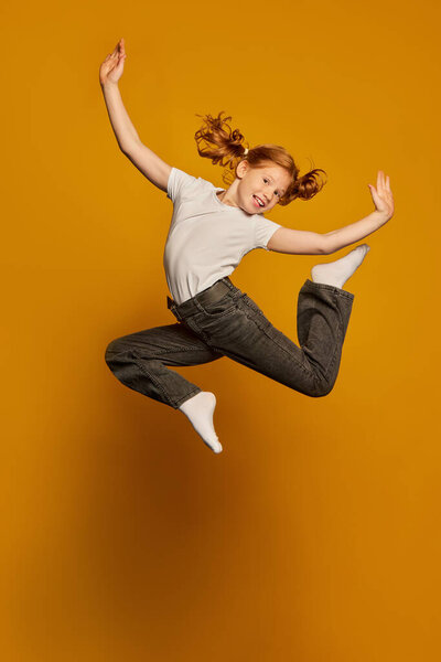Dance. Happy little girl, kid in white t-shirt and jeans jumping high isolated over yellow background. Kids fashion, emotions, carefree childhood, challenges, education concept