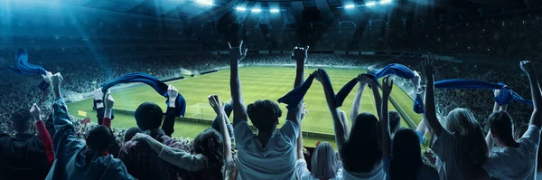 Back view of excited football, soccer fans cheering their team with blue scarfs at crowded stadium at evening time. Concept of sport, cup, world, team, event, competition