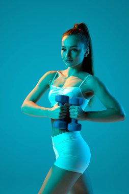 Beyaz spor giyim eğitimli genç bir kız. Dambılları mavi stüdyonun arka planında, neon ışıklarıyla izole edilmiş. Modern spor, aksiyon, hareket, gençlik konsepti. Fitness, hobi, sağlıklı yaşam tarzı