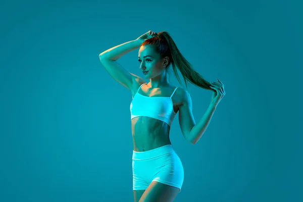 ネオンの光の中で青いスタジオの背景に隔離された白いスポーツウェア姿の若いスポーツウェアの女の子 筋肉質の体にフィット 現代のスポーツ アクション 若者の概念 フィットネス 健康的なライフスタイル — ストック写真