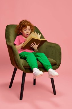 Şirin küçük bir kızın portresi, pembe arka planda izole edilmiş kıvırcık kızıl saçlı, duygusal kitap okuyan bir çocuk. Çocukluk kavramı, duygular, yaşam tarzı, moda, mutluluk. İlan için alanı kopyala
