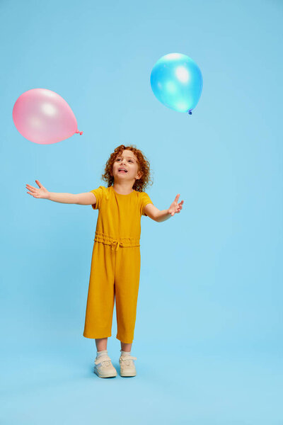 Portrait of cute little girl, child with curly red hair posing, catching balloons isolated over blue background. Concept of childhood, emotions, lifestyle, fashion, happiness. Copy space for ad