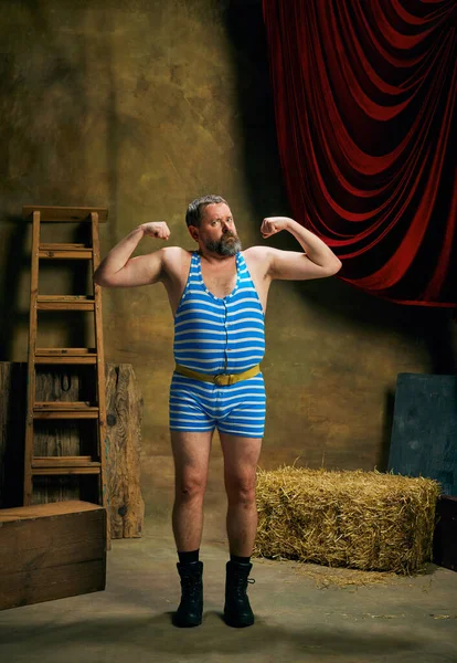 Strength. Mature man, retro circus strongman wearing striped sports swimsuit posing isolated over dark vintage circus backstage background. Art, 30s, 40s fashion style and inspiration