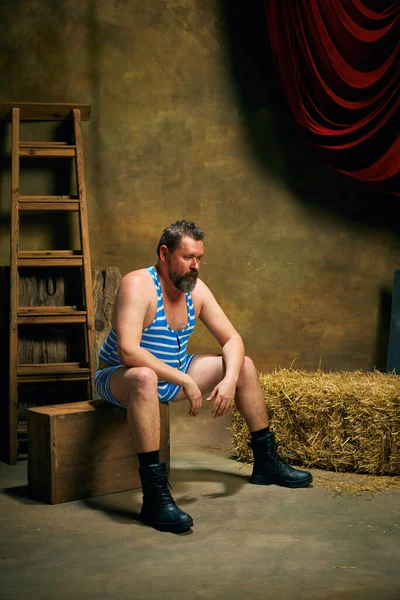 Mature man, retro circus strongman wearing striped sports swimsuit posing isolated over dark vintage circus backstage background. Art, 30s, 40s fashion style and inspiration