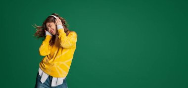 Genç güzel bir kızın portresi, duygusal olarak yeşil arka planda izole edilmiş kulaklıkla müzik dinliyor. Gençlik kavramı, güzellik, moda, yaşam tarzı, duygular, yüz ifadesi. Ad