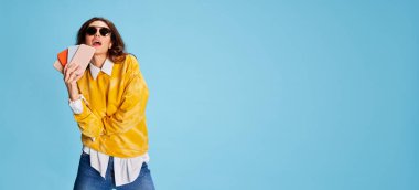 Mavi arka planda izole edilmiş telefonlarla poz veren güzel bir kızın portresi. Alışveriş, aletlerde büyük satışlar. Gençlik kavramı, güzellik, moda, yaşam tarzı, duygular, yüz ifadesi. Reklam. El ilanı