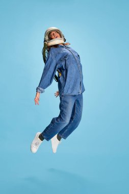 Üniforma içinde poz veren güzel bir kızın portresi ve mavi arka planda izole edilmiş astronot kaskı. Atlıyorum. Gençlik kavramı, güzellik, moda, yaşam tarzı, duygular, yüz ifadesi. Ad