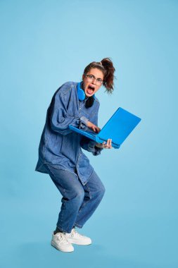 Portrait of young emotive girl, student posing, fastly typing on laptop isolated over blue background. Burning deadlines. Concept of youth, beauty, fashion, lifestyle, emotions, facial expression. Ad clipart