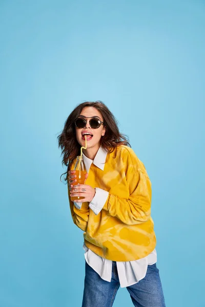 年轻美丽女孩的肖像是喝橙汁 戴着墨镜 与蓝色背景隔离 随意的 生活方式 面部表情的概念 — 图库照片