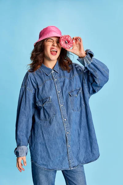 Mavi arka planda izole edilmiş pembe panamalı donutla poz veren genç duygusal kız portresi. Şekerler. Gençlik kavramı, güzellik, moda, yaşam tarzı, duygular, yüz ifadesi. Ad