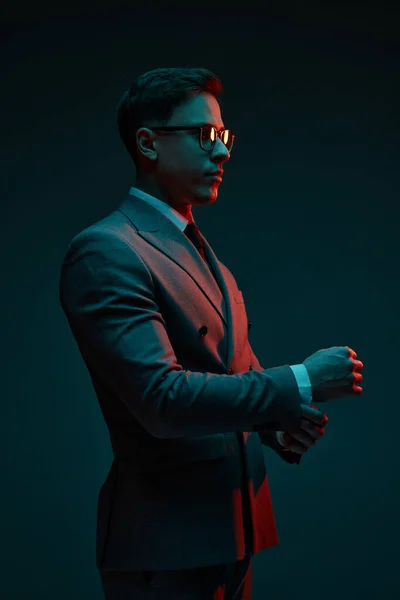 Fashionable serious young businessman in classic style suit adjusting his jacket sleeve isolated over dark background in neon light. Concept of business, fashion, style, modern lisfestyle.