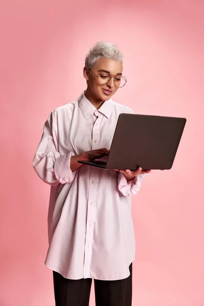 年轻时尚女性的肖像 身穿白衬衫 在笔记本电脑上打字 在粉色背景下独立工作 生活方式 面部表情的概念 — 图库照片