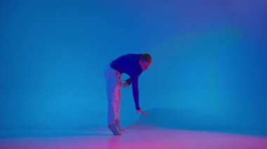 Genç, esnek, enerjik adam dans ediyor parlak spor kıyafetlerle deneysel dans ediyor mavi arka planda dans salonunda neon ışıkta. Modern gençlik kültürü, stil ve moda kavramı, eylem.