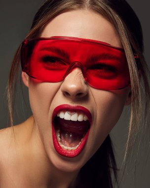Kırmızı gözlüklü, koyu gri arka planda bağıran duygusal genç kadının yakın plan portresi. Beyaz dişler ve kırmızı dudaklar estetik. Güzellik kavramı, moda, makyaj, dergi, duygular, reklam