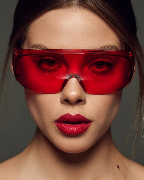 在深灰色背景下 身穿红色塑料眼镜 头戴红色口红的年轻漂亮女子的特写 化妆品治疗 情感和广告的概念 — 图库照片