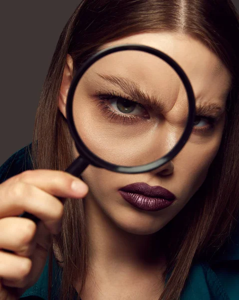 Close-up portrait of beautiful woman with dark lips posing, holding magnifying glass over dark grey background. Perfect eyebrows, lamination. Concept of beauty, fashion, magazine, emotions and ad.