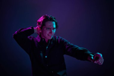 Siyah tişörtlü genç bir adamın portresi neon ışıkta koyu mor arka plan üzerinde gergin bir şekilde izole görünüyor. İnsan duyguları, yüz ifadesi, satış, reklam, moda ve güzellik kavramı