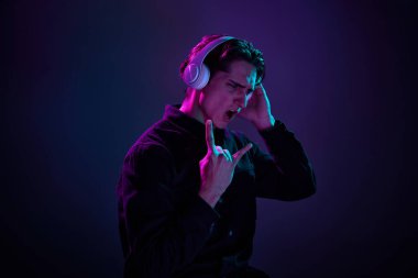 Siyah gömlekli, kulaklıkla poz veren, neon ışıkta koyu mor arka planda izole genç bir adam portresi. Rock müziği. İnsan duyguları kavramı, yüz ifadesi, satış, reklam, moda