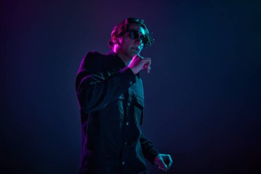 Gündelik siyah gömlekli ve modaya uygun güneş gözlüklü genç bir adamın portresi, neon ışıkta koyu mor arka planda izole bir şekilde poz veriyor. İnsan duyguları kavramı, yüz ifadesi, satış, reklam, moda