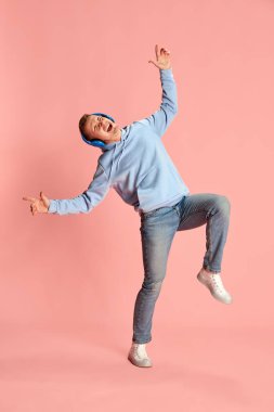 Kapüşonlu ve kot pantolonlu, kulaklıkla müzik dinleyen ve pembe arka planda dans eden genç duygusal bir adamın portresi. Gençlik kavramı, yaşam tarzı, müzik, moda, duygular, yüz ifadesi