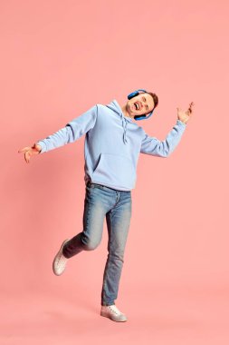 Kapüşonlu ve kot pantolonlu bir gencin portresi kulaklıkla müzik dinliyor ve pembe arka planda duygusal olarak izole bir şekilde şarkı söylüyor. Gençlik kavramı, yaşam tarzı, müzik, moda, duygular, yüz ifadesi