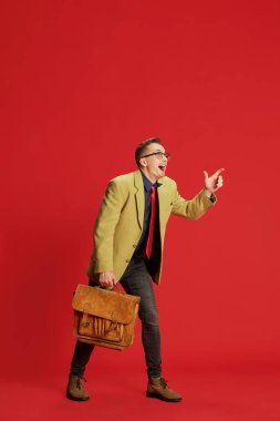 Kırmızı arka planda poz veren şık ceketli, gözlüklü ve antika çantalı bir gencin portresi. Yeni fikirler. Beyin fırtınası. Duygular, iş, meslek, hobi, yaşam tarzı, moda kavramı