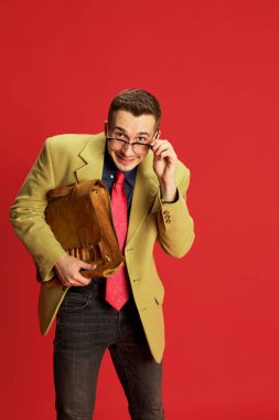 Kırmızı arka planda poz veren şık ceketli, gözlüklü ve antika çantalı bir gencin portresi. Yeni işçi, stajyer. Utangaç bir bakış. Duygular, iş, meslek, hobi, yaşam tarzı, moda kavramı