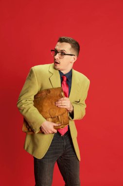 Şık ceketli, gözlüklü ve kırmızı arka planda ciddi bir yüzle poz veren klasik çantalı genç bir adam portresi. Duygular, iş, meslek, hobi, yaşam tarzı, moda kavramı