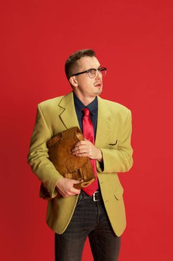 Şık ceketli, gözlüklü ve klasik çantalı genç bir adamın portresi kırmızı arka planda izole edilmiş sorgulama yüzüyle poz veriyor. Duygular, iş, meslek, hobi, yaşam tarzı, moda kavramı