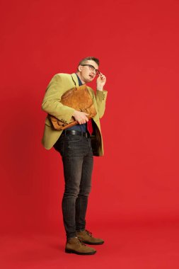 Kırmızı arka planda poz veren şık ceketli, gözlüklü ve antika çantalı bir gencin portresi. Yanlış anlaşılma. Duygular, iş, meslek, hobi, yaşam tarzı, moda kavramı