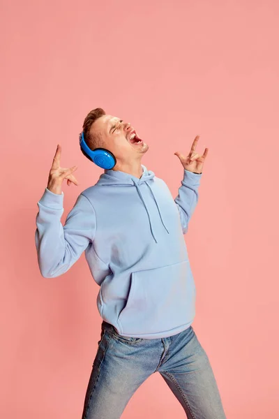 ピンクの背景の上に隔離されたヘッドフォンで音楽を聴くパーカーとジーンズの若い感情的な男の肖像 ライフスタイル カジュアルなファッション 表情の概念 — ストック写真