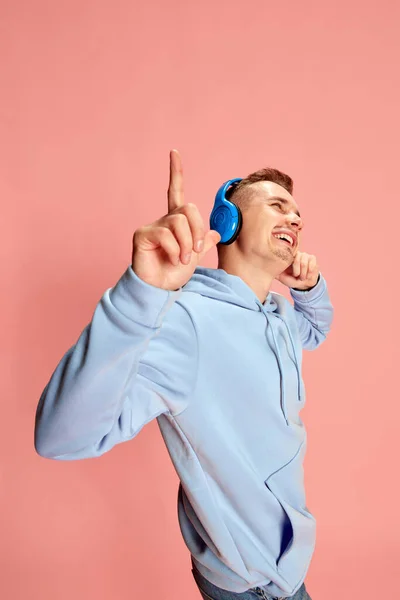 ピンクの背景の上に隔離されたヘッドフォンで音楽を聴く青いパーカーの若い陽気な男の肖像画 ライフスタイル カジュアルなファッション 表情の概念 — ストック写真