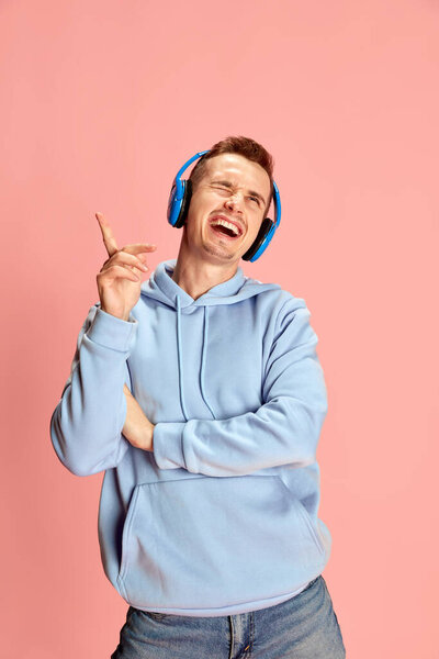 Portrait of young man in hoodie listening to music in headphones and emotionally singing isolated over pink background. Concept of youth, lifestyle, music, casual fashion, emotions, facial expression