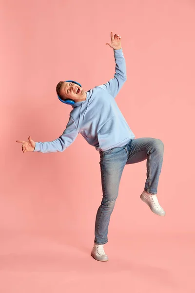 ヘッドフォンで音楽を聴き ピンクを背景に孤立して踊るパーカーとジーンズの若い感情的な男の肖像 ライフスタイル ファッション 表情の概念 — ストック写真