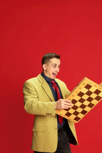 赤い背景の上にチェス盤でポーズをとるスタイリッシュなジャケットとメガネの若い男の肖像画 幸せと興奮 ビジネス ライフスタイル ファッションの概念 — ストック写真